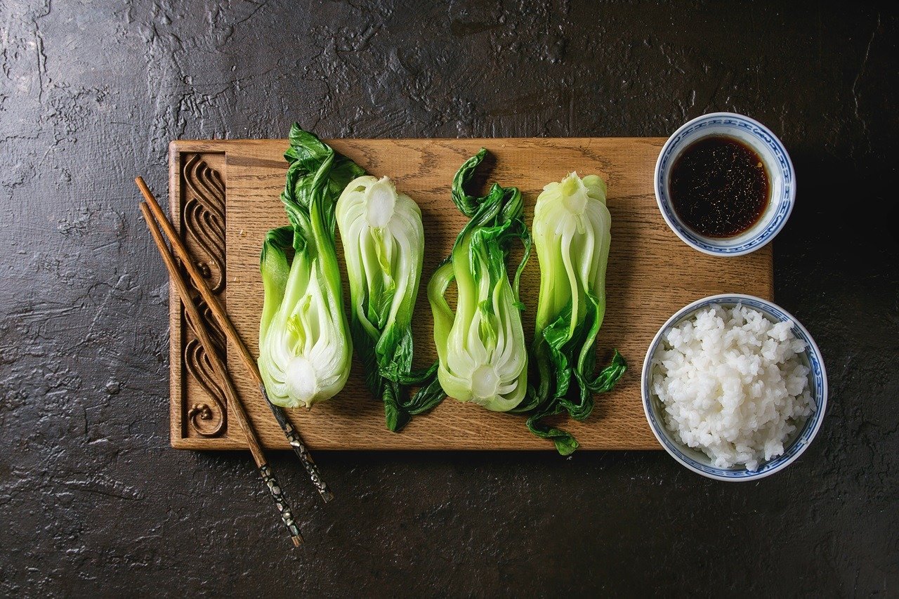 Stir fried bok choy or chinese cabbage with soy sauce and bowl of rice served on decorative wooden cutting board with chopsticks over dark texture background. Top view with space. Asian style dinner
